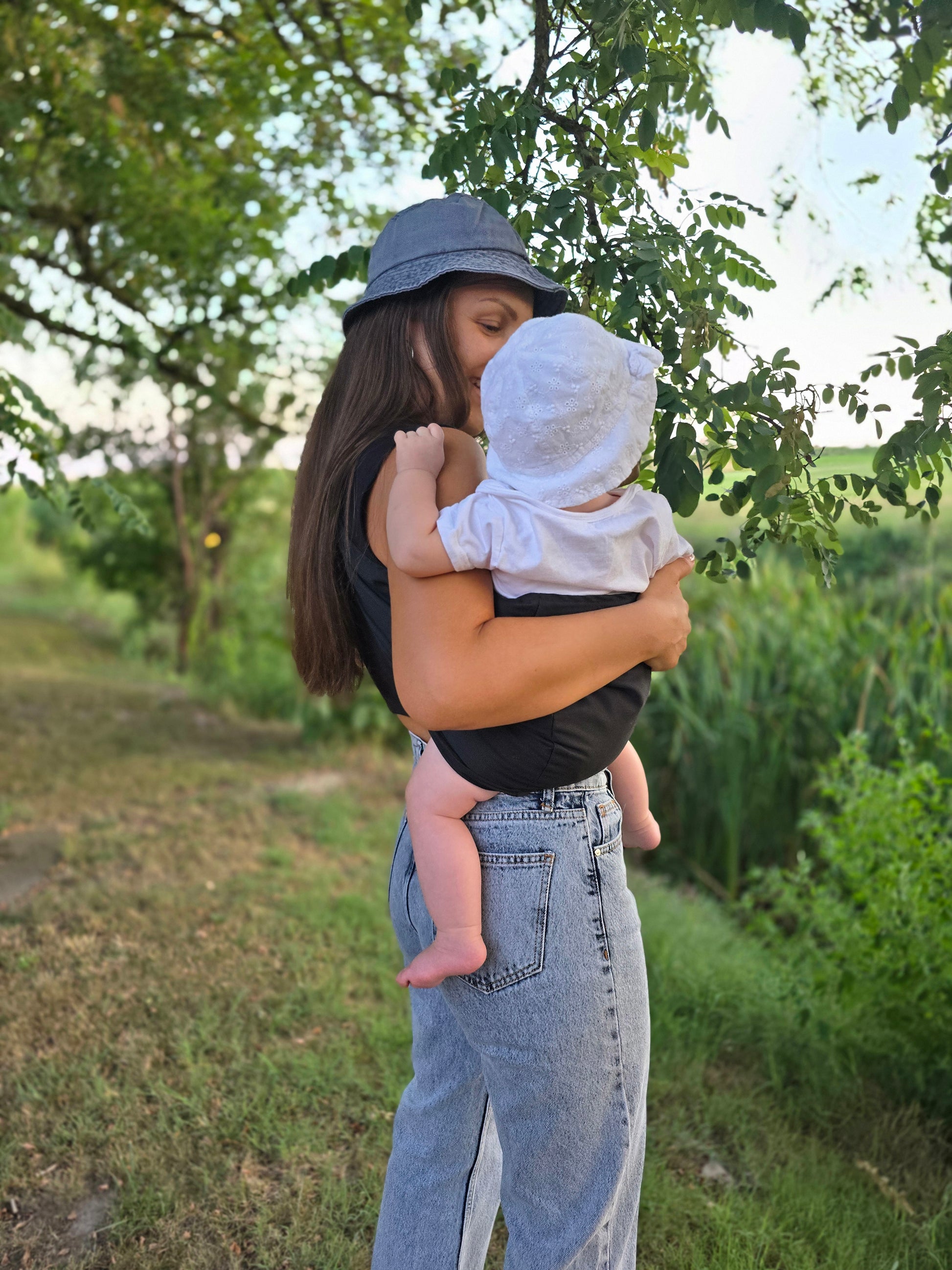 A mother wearing a gray bucket hat holding her baby in a Nest for Rest black toddler carrier outdoors. The scene is set in a lush green environment with trees and grass, capturing a tender moment between the mother and child as they enjoy nature together.