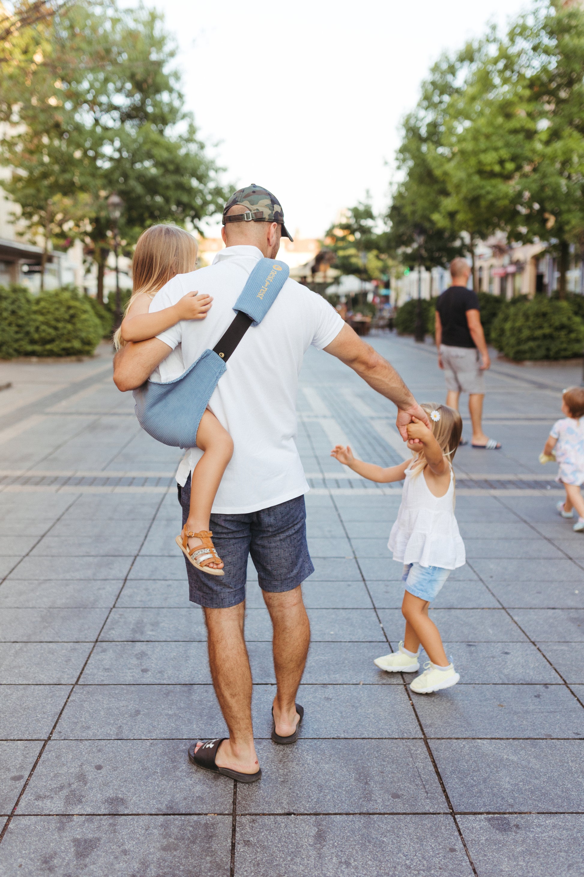 A father carrying his toddler in a Nest for Rest toddler carrier while holding hands with another child, walking down a busy pedestrian street. The carrier is designed for convenience and comfort, ideal for active parents on the go.