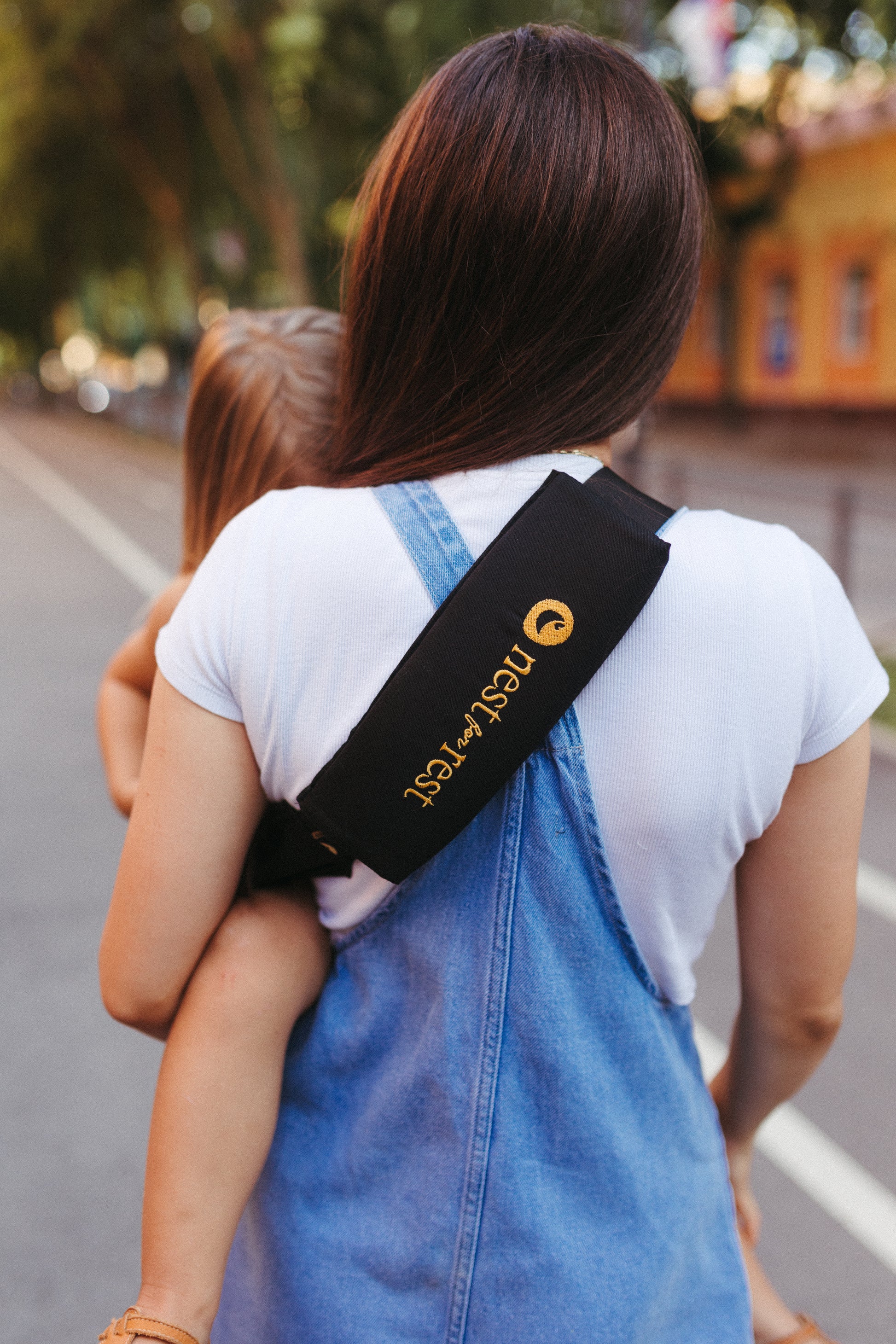 A mother wearing a black Nest for Rest toddler carrier with a golden logo, carrying her child on her back while walking outdoors. The carrier strap is securely positioned across the woman's shoulder, and the scene captures a peaceful, tree-lined street in the background.