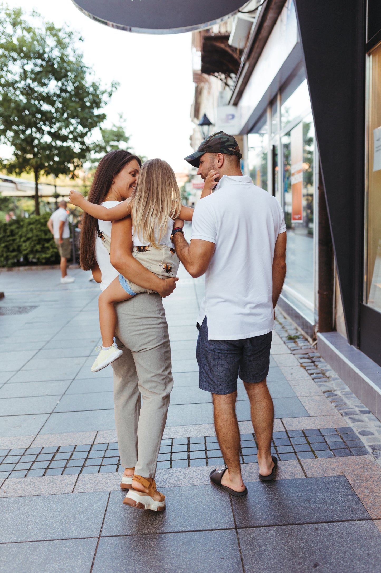 A family walking together in an urban setting. The mother, carrying a young child in a Nest for Rest toddler carrier with a bumblebee pattern, is smiling at her partner. The father, wearing a white polo and shorts, looks at the child, creating a warm family moment. The backdrop includes a modern city street with storefronts and greenery