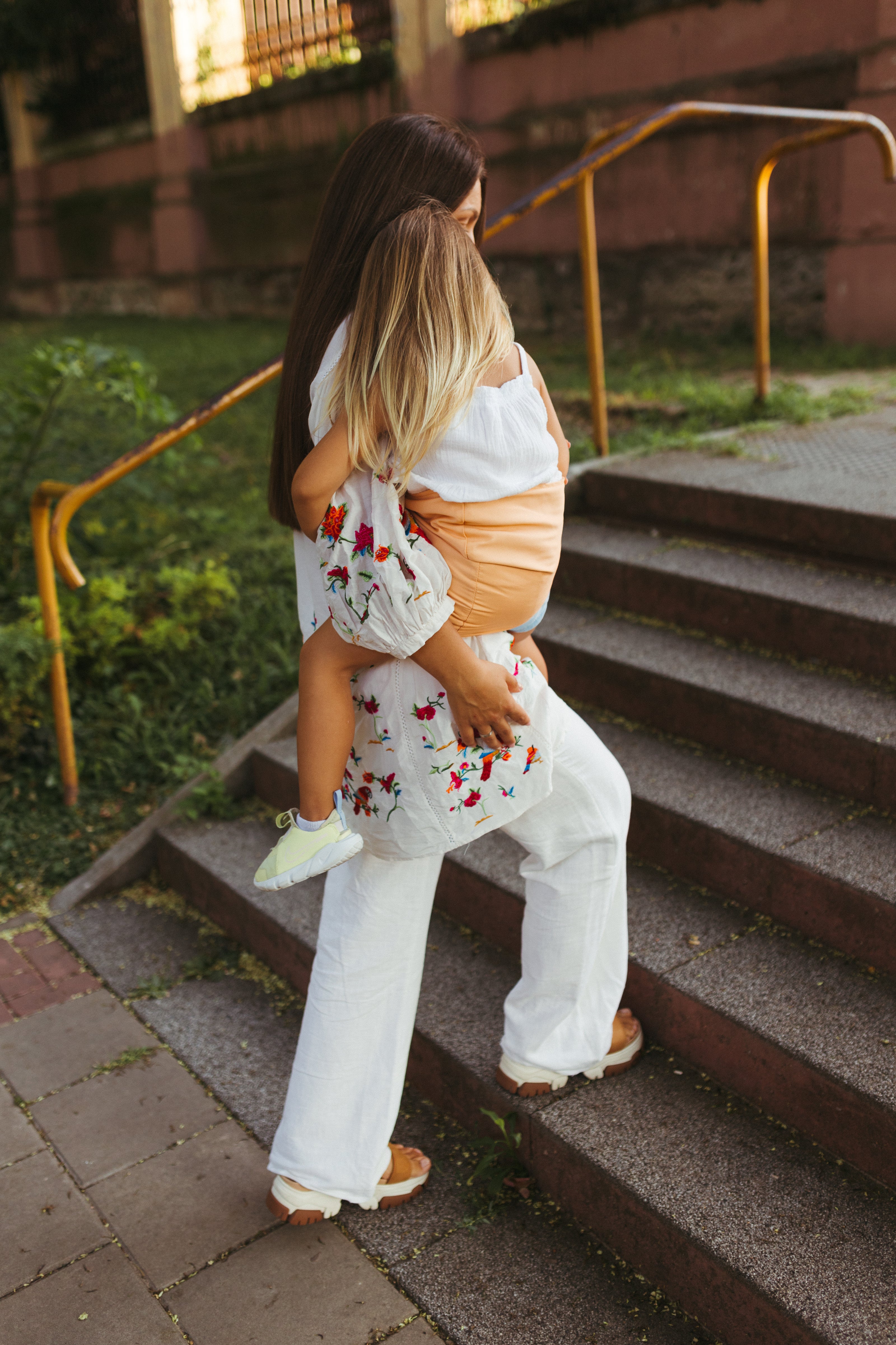 Mother carrying her child in a peach-colored 'Nest for Rest' toddler carrier while walking up a set of outdoor stairs. The scene captures the secure and ergonomic design of the carrier, providing comfort and support for both the parent and the child. The mother is dressed in a stylish white outfit with floral embroidery, and the child wears a white dress and neon green sneakers, adding a vibrant touch to the image.