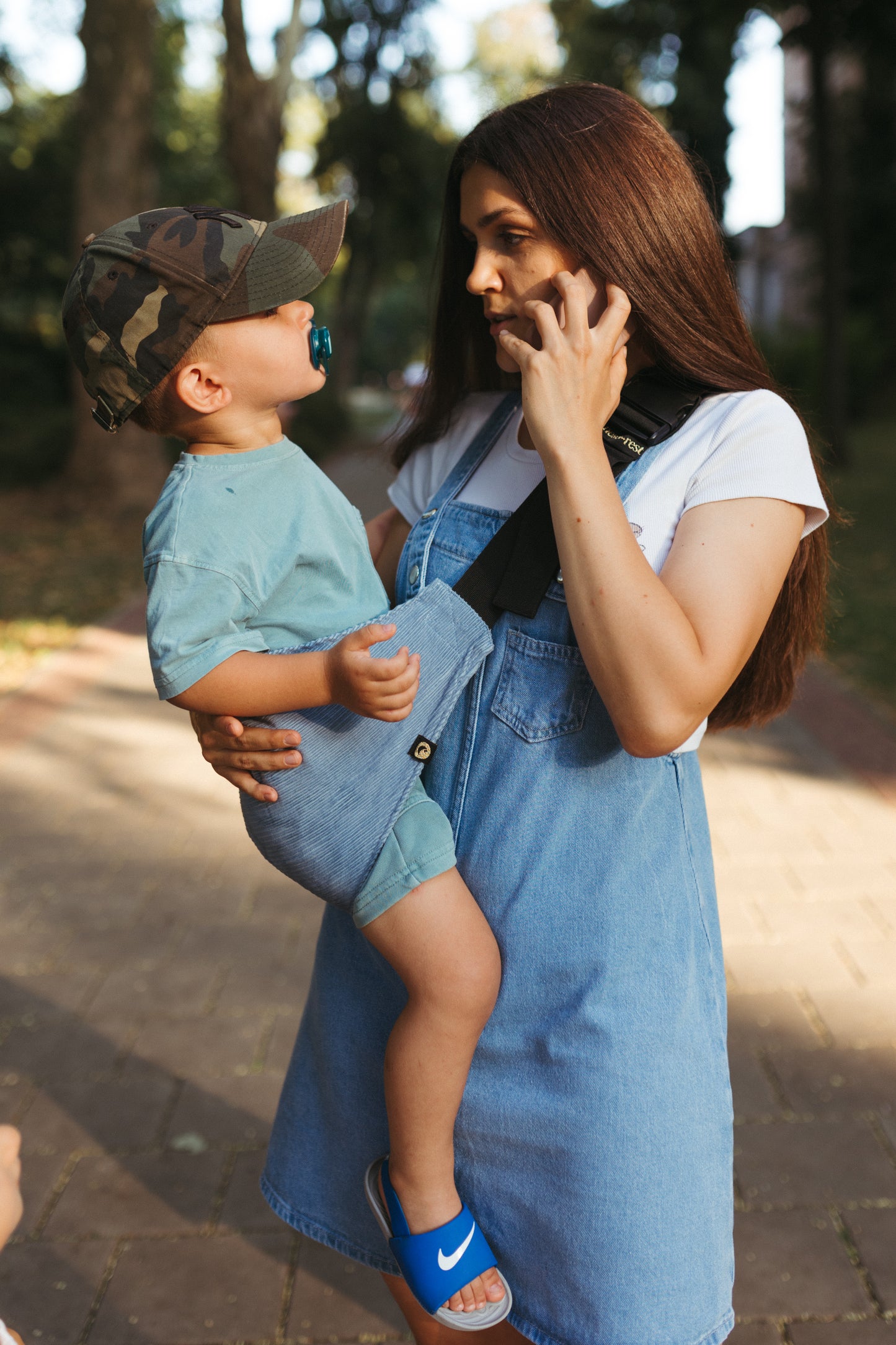 A woman in a denim dress carrying her toddler in a Nest for Rest carrier, standing outdoors in a park or garden with trees in the background.