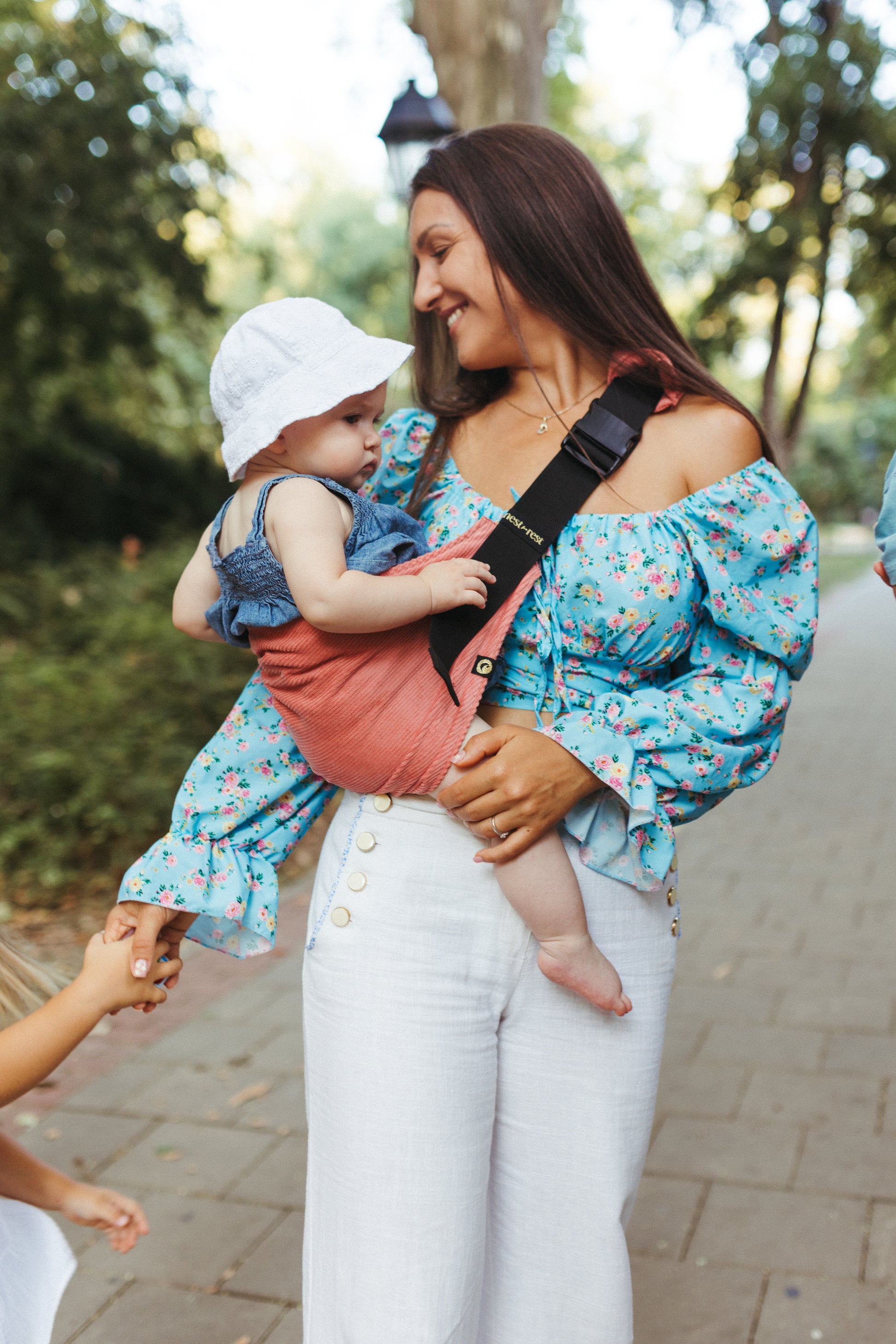 Mother wearing a floral blouse, carrying her baby in a Nest for Rest toddler carrier. The baby is secure and comfortable in the carrier, ideal for outdoor activities where a stroller isn't practical. The Nest for Rest carrier offers hands-free convenience, making it perfect for busy parents on the go.