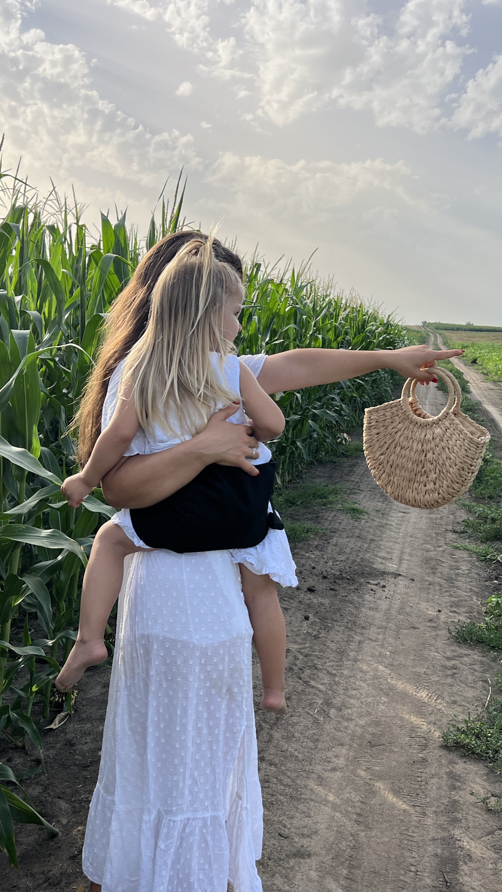 A mother and daughter walk along a dirt path beside a tall cornfield under a partly cloudy sky. The mother, dressed in a flowing white dress, carries her daughter in a black Nest for Rest toddler carrier. The child points forward, and the mother holds a straw handbag in her other hand. The scene captures a tranquil moment in nature, emphasizing the ease and comfort of using the Nest for Rest black toddler carrier during outdoor adventures.