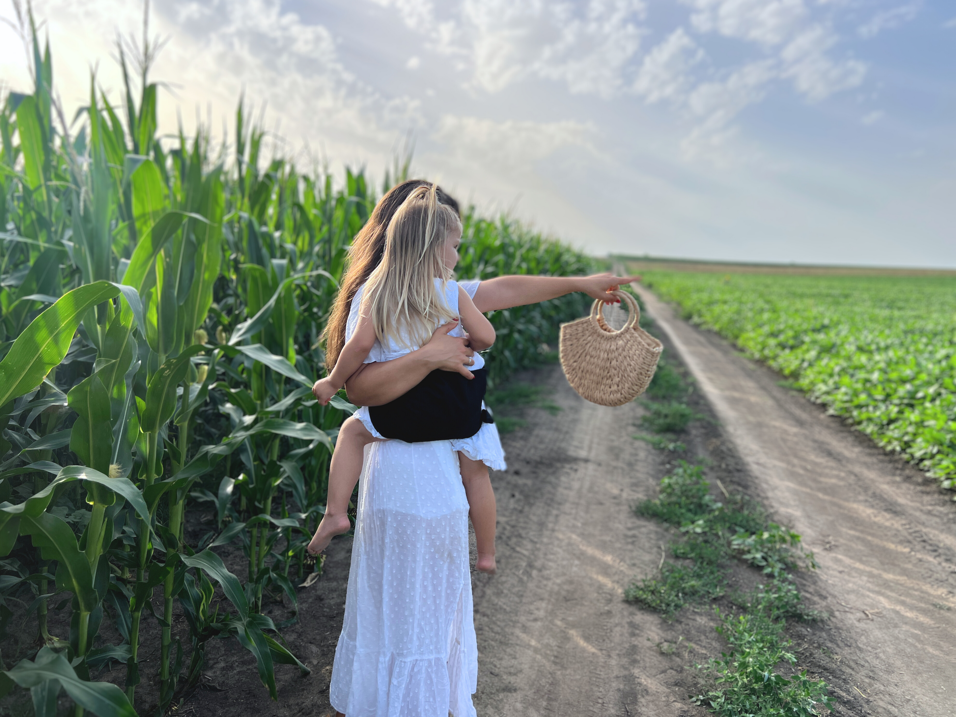 A mother and daughter walk along a dirt path beside a tall cornfield under a partly cloudy sky. The mother, dressed in a flowing white dress, carries her daughter in a black Nest for Rest toddler carrier. The child points forward, and the mother holds a straw handbag in her other hand. The scene captures a tranquil moment in nature, emphasizing the ease and comfort of using the Nest for Rest black toddler carrier during outdoor adventures.